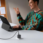 A man in a green, brown, and black patterned sweater sits at a white desk using a black laptop. In the foreground, a black power adapter sits on a small white stand, connected by a black cable.