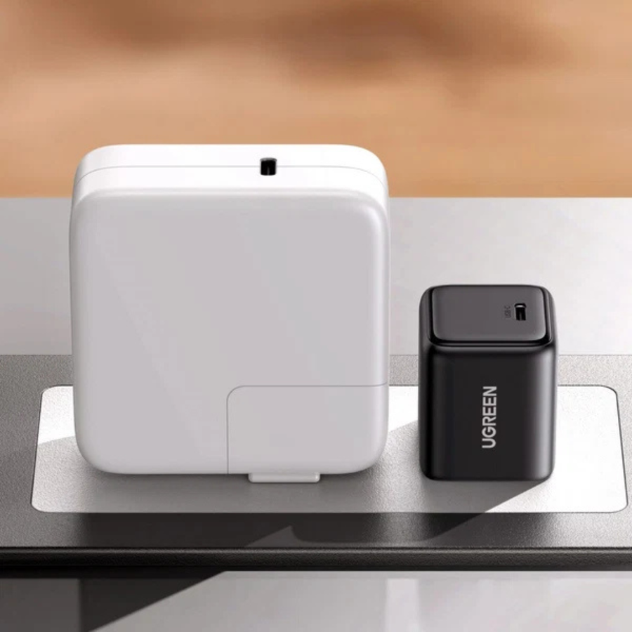 A white square electronic device with rounded corners stands next to a black UGREEN cube-shaped charger on a reflective dark surface against a warm beige blurred background.