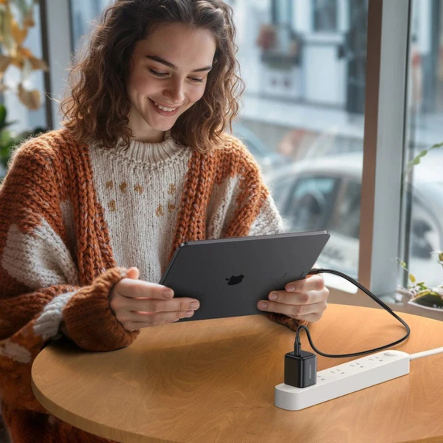 A woman in a chunky knit cardigan smiles while holding a tablet, with a power adapter plugged into a white power strip on a wooden table in a bright indoor setting.