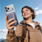 A woman outdoors wearing a brown fleece-lined zip jacket and backpack, holding a silver smartphone with a large lavender-gray magnetic case attached to the back. The background shows a bright blue sky with a few clouds.