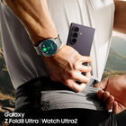 A close-up lifestyle shot of a person wearing a Samsung Galaxy Z Fold8 Ultra smartphone and a Samsung Watch Ultra2. The person is shown from the chest down, wearing a white tank top and black shorts, with the phone tucked into their waistband.