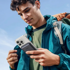 A man outdoors wearing a teal jacket and backpack, holding a smartphone with a black MagSafe-style magnetic case attached, against a blue sky background.