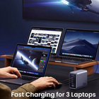A portable 300W power bank in a metallic silver-gray case is shown charging three laptops on a desk. A person's hands and arms are visible using the laptops, and the scene is set in a dark, modern workspace with a wooden monitor stand and a dark blue wall.