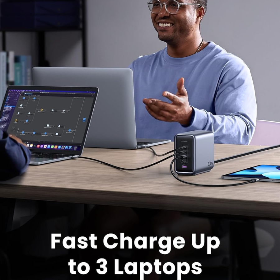 A man sits at a desk using laptops and a tablet, with a dark gray multi-port charger in the foreground. Text at the bottom reads 'Fast Charge Up to 3 Laptops'.