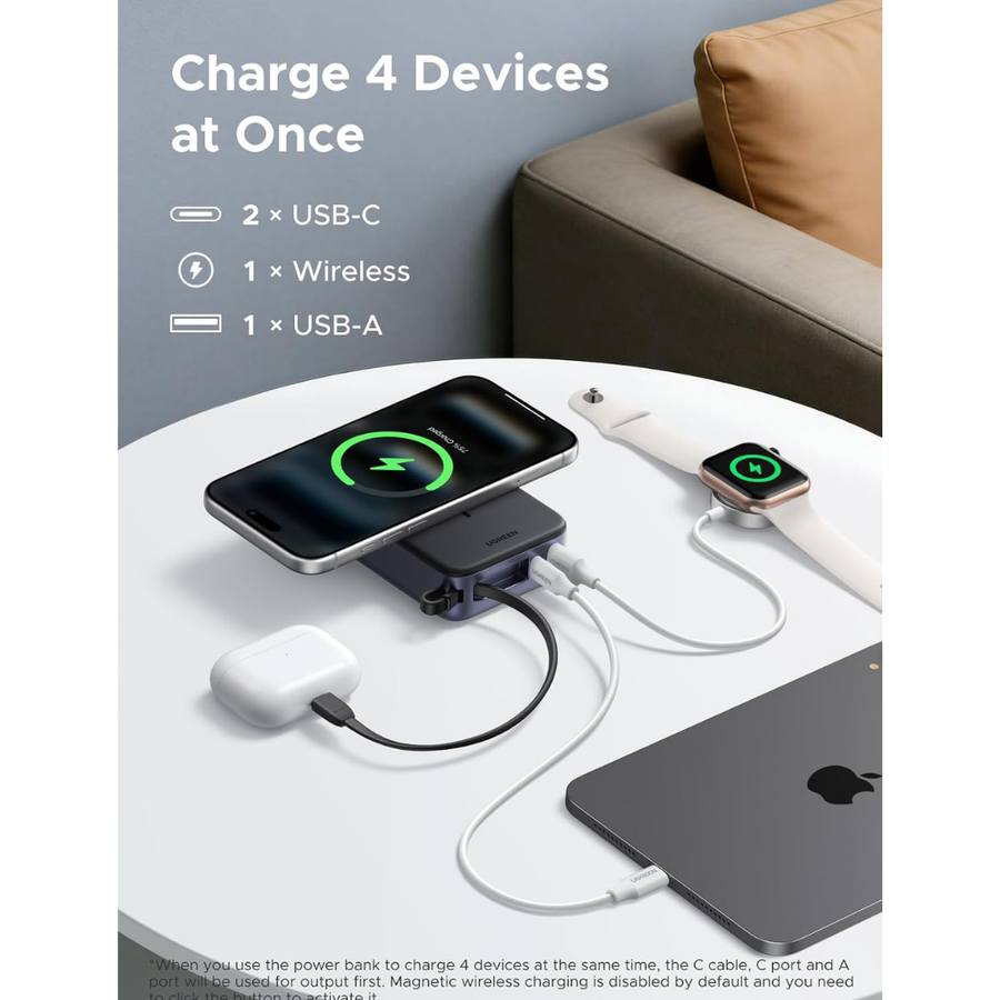 A portable multi-device wireless charging power bank shown on a white round table in a living room setting, charging a smartphone, AirPods, an Apple Watch, and a laptop. The image includes text highlighting its capability to charge 4 devices at once.